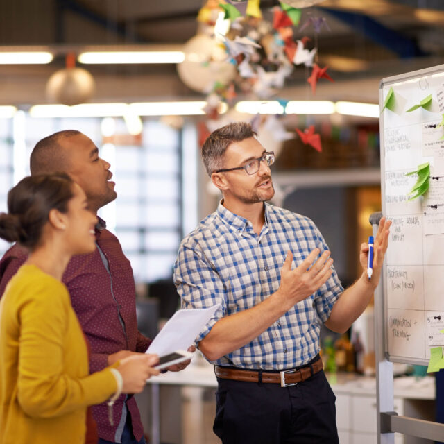 A global company strategizing, with a group of people standing around a whiteboard in an office.